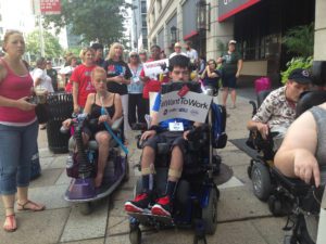 Young man sitting in wheelchair participates in street march, holding a sign that reads: “I Want to Work.”