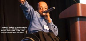 Chaz Kellem, a black man holding a microphone and using a wheelchair, wears a blue button down and black dress pants.