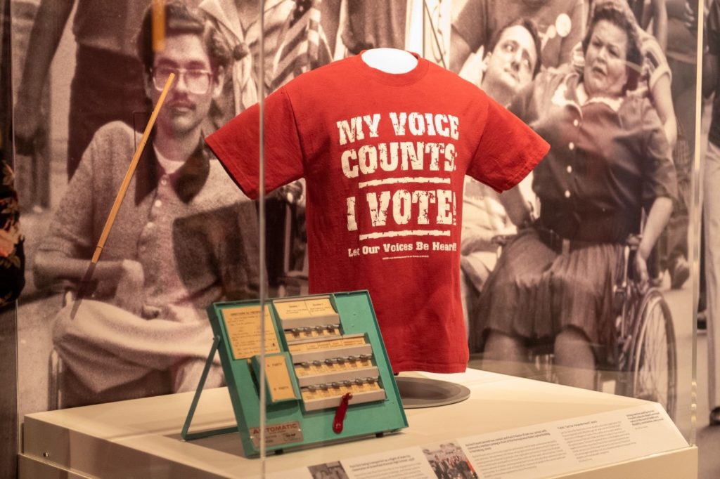 Artifacts from Paul Dick and Rachel Freund on display in American Democracy: A Great Leap of Faith. From left to right: dowel rod used by Paul to vote, voting machine used by Rachel to train voters, and Let Our Voices Be Heard t-shirt worn by Rachel. The backdrop is a black and white image of disability rights activists including Eileen Shackleton and Ray Smith marching in a parade. Image Source: Senator John Heinz History Center