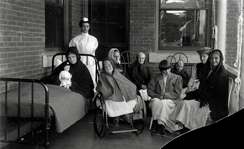 Black and white image of institutionalized women in the company of a nurse on a porch at Polk State Center, c. 1900s-1920s.