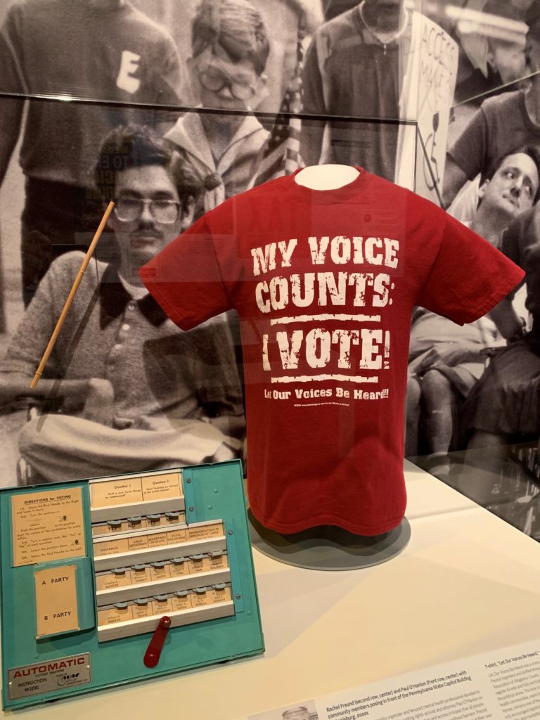 Photo shows detail from the disability rights segment of the American Democracy exhibit. On the right is a red T-shirt with the words “My Voice Counts: I Vote” in large white letters. On the left is a voting machine used in an advocacy effort to educate voters with disabilities.
