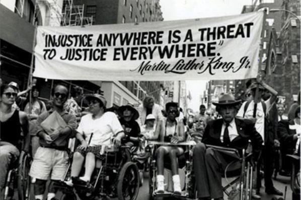 Photo shows a disability rights march in on a city street. Above the marchers is a banner with a quote from Martin Luther King that reads: “Injustice anywhere is a threat to justice everywhere.”