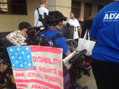 The back view of a man using a wheelchair, a sign on the back of the chair reads disability rights are civil rights.