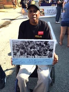 Milt Henderson using his wheelchair, with a sign that says Because They Marched