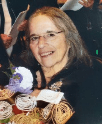 A recent photo of Judy Barricella. She is wearing glasses, smiling, and holding a bouquet of flowers.