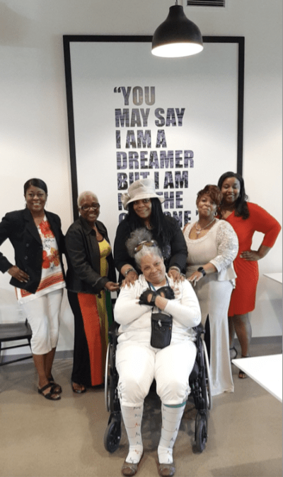 Andrea, using her wheelchair, standing with a group of five other black women in front of a sign with a John Lennon quote "You may say I'm a Dreamer, but I'm not the Only One."