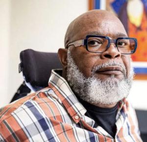 Theo Braddy head shot - a black man with a bald head, salt and pepper full beard, wearing eyeglasses, sitting in a wheelchair wearing a orange, blue and white plaid dress shirt