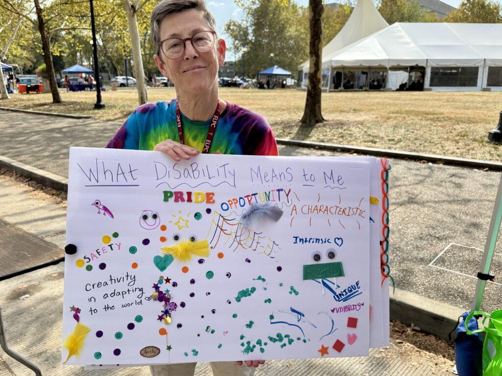 A different poster entitled What Disability Pride means to me. A woman with glasses and short hair, wearing a rainbow tie dyed shirt is holding the poster.