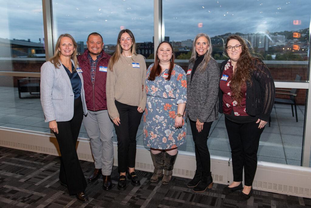 Five women and one man pose for a photo in front of the large windows.