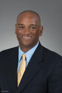 Curtis Pride head shot. He is a Black man with closely shaved hair, a big smile, using hear aids, and wearing a black suit with a blue shirt and yellow necktie.